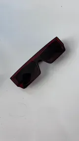 Lentes de sol de montura gruesa y forma rectangular, color carey oscuro o marrón rojizo. Los cristales son oscuros y tintados.