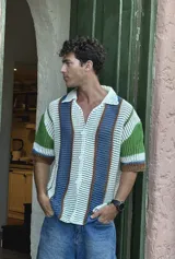 Camisa de manga corta tejida a crochet, con cuello polo y botones frontales. Presenta un diseño de rayas verticales en blanco, azul, verde y marrón.