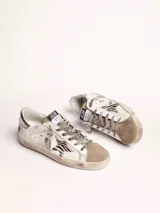 Zapatillas urbanas Golden Goose modelo Super-Star, de cuero blanco con efecto desgastado. Presentan una estrella de animal print en el lateral, talón plateado y cordones grises con inscripciones.