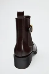 Botín chelsea color marrón oscuro de cuero sintético con elásticos laterales, hebilla decorativa dorada y tirador trasero.
