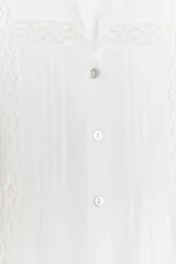 Camisa blanca de cuello alto y manga larga, con detalle de entredós combinados a tono y cierre frontal con botones.
