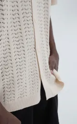 Camisa de tejido de punto color beige, manga corta, con cuello polo y cierre frontal de botones. Presenta un calce regular y un patrón calado en zigzag.