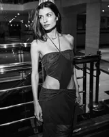 Vestido largo negro, ajustado al cuerpo, con escote strapless y tirantes finos cruzados en el cuello. Presenta recortes laterales pronunciados en la cintura y un nudo fruncido en la parte inferior frontal.