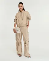 Pantalón cargo color beige confeccionado en gabardina gastada de lino y algodón. Silueta de pierna recta ancha, con cartera con cierre metálico y broche níquel en el cruce de la cintura. Tiene bolsillos inclinados en los laterales y bolsillos cargo con tapa y broches metálicos a la altura de las rodillas, de los cuales salen unas tiras que se ajustan mediante hebillas metálicas para modificar la silueta del pantalón. En la espalda tiene pinzas y un bolsillo plaque con cierre metálico en boca. Es tiro medio.