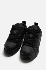 Zapatilla deportiva tipo skate color negro. Parte superior fabricada en combinación de piezas y materiales. Detalle de cordones maxi y suela con acabado irregular.
