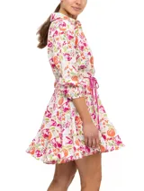 Vestido camisero corto con estampado floral en tonos beige, celeste y verde. Tiene cuello camisero, mangas 3/4 con puños elásticos, cierre frontal con botones y cinturón fino con borlas.
