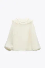 Camisa color crudo confeccionada en tejido con lyocell. Presenta escote en pico, mangas largas abullonadas con elástico en los puños, volantes frontales y cierre frontal con botones.