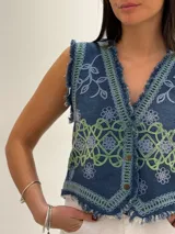 Chaleco corto de denim azul con bordados florales en blanco y marrón, detalle desflecado en escote, sisas y ruedo. Escote en V y cierre frontal con botones. Estilo boho chic.