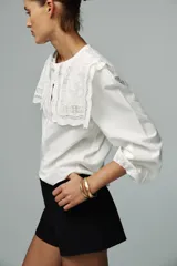 Camisa blanca de popelín de algodón con cuello redondo con tejido en superposición a tono y aplicación de bordados. Manga larga acabada en elástico y cierre frontal con botones ocultos por sopata.