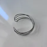 Brazalete abierto de metal plateado, estilo cuff, compuesto por múltiples alambres delgados que se cruzan y entrelazan de forma abstracta.