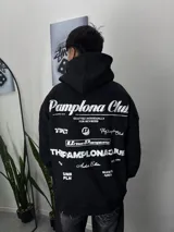Buzo negro con capucha y cordones ajustables. Estampado en la espalda con el texto "Pamplona Club" en letras grandes y otros textos y logos más pequeños.