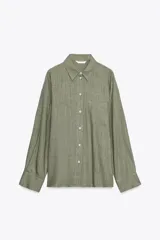 Camisa verde oliva amplia de lino, con cuello solapa, manga larga, bolsillo de plastrón en el delantero, bajo asimétrico y cierre frontal con botones.