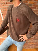 Sweater de tejido de punto color beige, con cuello redondo y manga raglán. Presenta un pequeño bordado en hilo rojo en el pecho.