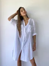 Vestido camisero blanco de corte holgado, con cuello mao, cierre frontal con botones, mangas 3/4 con puños abotonados, dos bolsillos de parche en el pecho y aberturas laterales en el ruedo.