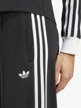 Pantalón de sudadera Adidas de corte ancho, color negro con tres rayas blancas verticales en los laterales y logo bordado en la parte superior. Confeccionado en tejido de felpa francesa.