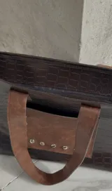 Bolso tote negro con textura símil cuero de cocodrilo, bolsillo marrón con tachas plateadas en el frente y asas de cuero marrón.