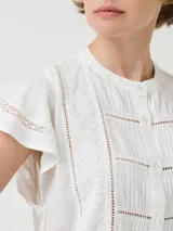 Blusa blanca de lino orgánico y viscosa, con cuello redondo, cierre frontal con botones, mangas cortas con volados y broderie con calados en el frente.