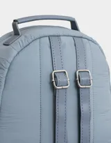 Mochila color crema de tela acolchada con pespuntes verticales. Tiene un bolsillo frontal con cierre, doble cierre superior, manija superior, correas regulables y bolsillos laterales. Incluye un charm colgante de corazón.