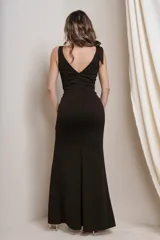 Vestido largo de fiesta negro, con escote en V profundo, breteles finos y lazo decorativo en el hombro. Tiene corte ajustado al cuerpo y abertura alta en la pierna.
