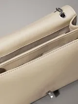 Cartera tipo sobre de piel sintética color beige claro, con solapa frontal y herrajes plateados. Presenta el monograma 'CK' en metal en la parte frontal y una correa de cadena convertible que permite usarla al hombro o cruzada.
