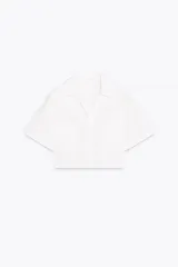 Camisa blanca de popelín con cuello solapa, escote pico y manga corta. Tiene bolsillos delanteros de plastrón y cierre frontal con botones.