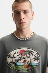Camiseta gris de corte recto con cuello redondo y mangas cortas. Estampado ovalado con palmeras, sol y texto "Coconut Amora. Maui Offers Paradise".