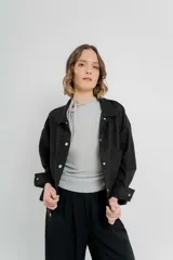 Chaqueta color beige de corte recto, estilo camisa, confeccionada en gamuza con elastano. Presenta cuello camisero, cierre frontal con botones metálicos, bolsillos grandes en el frente y charreteras en los hombros.