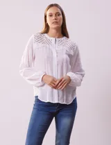 Blusa blanca de manga larga con cuello redondo y abertura central con botones. Presenta detalle de broderie en la parte superior delantera y terminación elastizada en los puños.