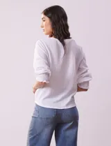 Blusa blanca de tejido texturado con cuello redondo, escote en V con aplique metálico y mangas 3/4 con puños elásticos. El ruedo se ata con un nudo.