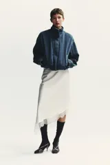 Chaqueta corta de denim azul con cuello alto, manga larga con hombros caídos, bolsillos laterales, bajo con elástico y cierre frontal con cremallera y botones ocultos.