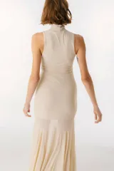 Vestido largo color beige, con cuello alto y sin mangas. La parte superior es ajustada al cuerpo y la falda es amplia y fluida, con paneles traslúcidos.