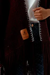 Ruana tejida a mano en lana merino 100%, de color marrón con detalles de costura en rojo en los bordes y terminación de flecos largos en el ruedo. Presenta una silueta amplia y caída natural, confeccionada artesanalmente por Manos del Uruguay.
