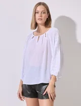 Blusa de gasa color blanco con cuello redondo y aberturas en V, detalles en puntilla y puño fruncido en las mangas.