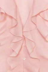 Camisa color rosa pálido confeccionada en tejido ligero con lyocell. Presenta escote en V, cierre frontal con botones, mangas largas abullonadas con puños elásticos y detalle de volantes en el frente.
