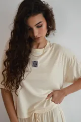 Remera de manga corta color crema, con textura acanalada y cuello redondo. Presenta un pequeño parche bordado con un diseño floral o geométrico en el pecho.