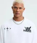 Remera blanca de algodón, corte oversized, con cuello redondo y mangas cortas. Presenta un estampado frontal con un ángel y el texto "PARADISE" en negro, y un pequeño gráfico de estrella/brújula en el lado izquierdo del pecho.