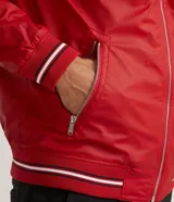 Chaqueta bomber roja de poliéster engomado, con cuello, puños y cintura acanalados a rayas blancas y azul marino. Tiene cierre frontal con cremallera y bolsillos laterales con cremallera.