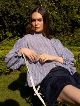 Camisa verde con rayas verticales blancas y bordados de flores. Tiene cuello mao, mangas largas abullonadas con puños elásticos y cierre frontal con botones de nácar.