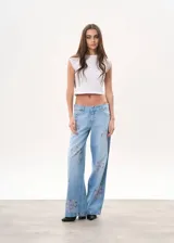 Pantalón de jean celeste de tiro bajo, corte wide leg y apliques de strass con forma de rosa en ambas piernas.