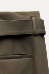 Pantalón de tiro medio color verde oliva, con cintura con trabillas, bolsillos laterales ocultos, cinturón ajustable con hebilla metálica, pinzas delanteras y cierre frontal con cremallera, botón y gancho metálico.