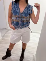 Chaleco corto de denim azul con bordados florales en blanco y marrón, detalle desflecado en escote, sisas y ruedo. Escote en V y cierre frontal con botones. Estilo boho chic.
