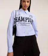 Camisa de manga larga, cuello camisero, abotonada, con rayas verticales finas celestes y blancas, y estampado grande en negro con la palabra "CHAMPION".