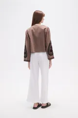 Chaqueta corta de algodón color marrón topo, sin cuello, con bordados florales y de estilo paisley en negro. Presenta un borde festoneado y mangas largas.