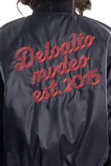 Campera bomber negra de tela satinada con cierre frontal, cuello redondo y puños y cintura elastizados. Tiene un pequeño logo bordado en el pecho y un bolsillo con cierre en la manga. Es reversible.