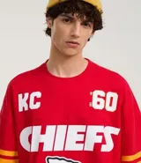 Remera roja de algodón con estampado del logo de los Kansas City Chiefs, el nombre del equipo y el número 60 en blanco. Tiene mangas cortas con rayas amarillas y rojas.
