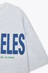 Camiseta gris jaspeado de corte amplio, cuello redondo y mangas cortas. Estampado frontal con la frase "Los Angeles 8500 Beverly Blvd, Los Angeles, CA 90048" en color azul.