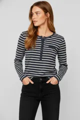 Remera de manga larga con cuello redondo y abertura frontal con botones. Presenta un patrón de rayas horizontales blancas y negras, y un pequeño bolsillo negro en el pecho.