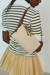 Bandolera rectangular de tela color beige con textura tipo canasta y detalles en cuero marrón. Tiene cierre superior con cremallera, correa larga regulable con hebilla y forro interior.