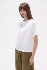Remera blanca de manga corta y cuello redondo, con un bordado floral en tonos azules en la zona superior izquierda del pecho.