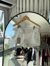 Short blanco de cuero sintético con cinturón con hebilla metálica y detalles de ojalillos metálicos. Presenta cordones cruzados en los laterales.